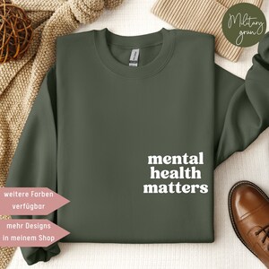 Könnte beinhalten: Ein dunkelgrüner Pullover mit dem Text "mental health matters" in weißen Buchstaben. Der Pullover liegt auf einem weißen Hintergrund mit einer braunen Decke und einem Paar braunen Schuhen.