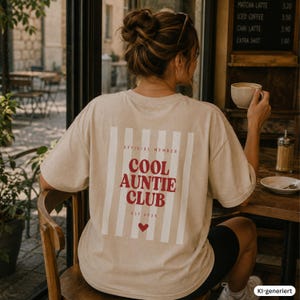 Könnte beinhalten: Ein beiges Kurzarm-T-Shirt mit einem Rückendruck aus vertikalen weißen Streifen. Der Text "OFFICIAL MEMBER COOL AUNTIE CLUB EST 2025" ist in roter Schrift gehalten, ergänzt durch ein kleines rotes Herzsymbol unter dem Text.
