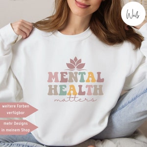 Könnte beinhalten: Weißer Pullover mit dem Text "Mental Health Matters" in bunter Retro-Schrift. Über dem Text befindet sich eine rosa Lotusblume. Der Text "weitere Farben verfügbar" und "mehr Designs in meinem Shop" befindet sich in der linken unteren Ecke des Bildes.