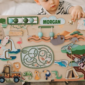 Op de afbeelding: Een houten activiteitenbord met een jungle thema. Het bord heeft verschillende activiteiten, waaronder een kralenlabyrint, een vormensorteerder, een draaiwiel en een schuifpuzzel. Het bord is gepersonaliseerd met de naam "MORGAN".
