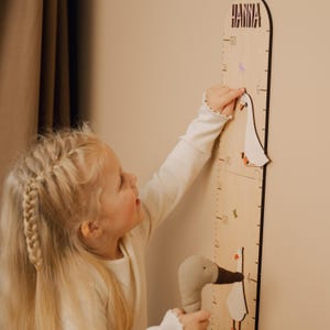 May include: Wooden growth chart with the name "HANNA" at the top. The chart features a goose design and measurements in inches. A child is pointing at the chart with a stuffed goose toy in their other hand.