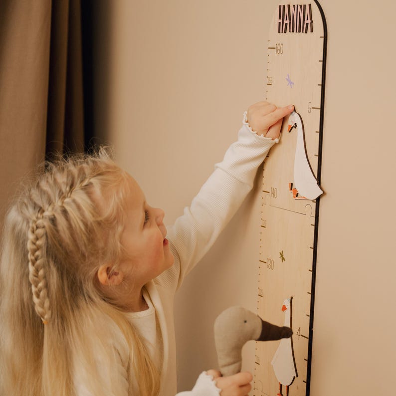 May include: Wooden growth chart with the name "HANNA" at the top. The chart features a goose design and measurements in inches. A child is pointing at the chart with a stuffed goose toy in their other hand.