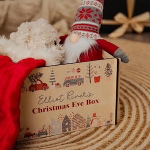 Op de afbeelding: Een kerstavonddoos met de tekst "Elliot River's Christmas Eve Box" en feestelijke illustraties. De doos bevat een witte pluchen hond en een kabouter met een rode en grijze hoed. Een rode kous is zichtbaar.