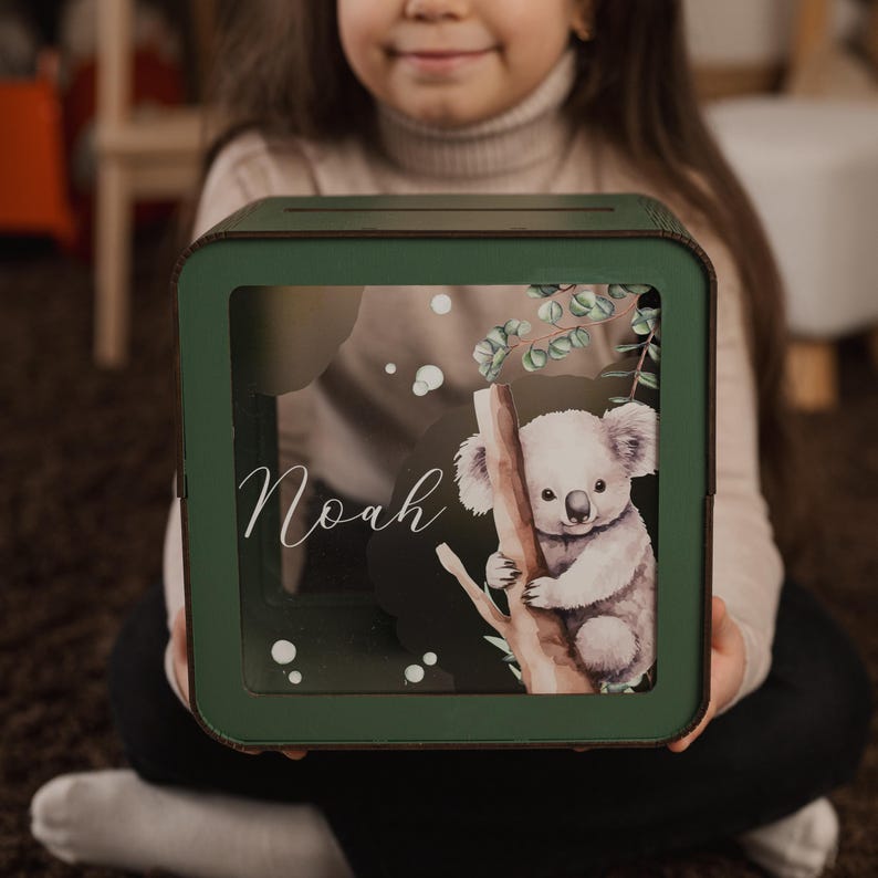 May include: Green wooden piggy bank with a clear front panel featuring a watercolor illustration of a koala holding onto a tree branch. The name "Noah" is written in white cursive lettering on the front of the bank.