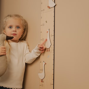 May include: A wooden growth chart with goose illustrations and centimeter markings. A young child points at the chart, holding a goose plush toy. The chart measures up to 140 cm, with goose cutouts at various heights.