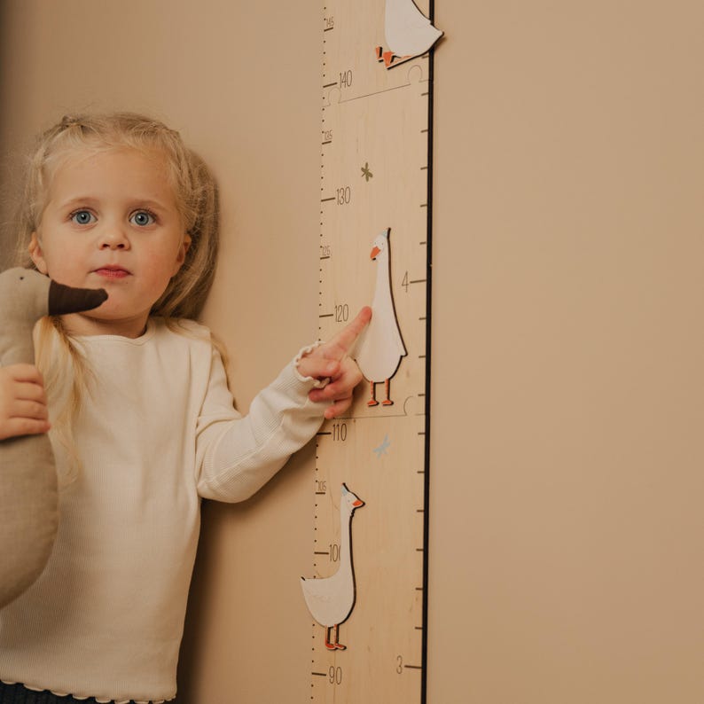 May include: A wooden growth chart with goose illustrations and centimeter markings. A young child points at the chart, holding a goose plush toy. The chart measures up to 140 cm, with goose cutouts at various heights.