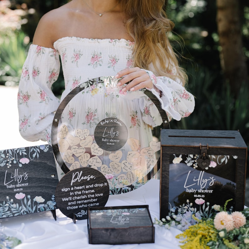 May include: A wooden box with a glass front and a floral design. The box has a sign that says "Lilly's Baby Shower" and a date. The box is sitting on a table with other decorations.