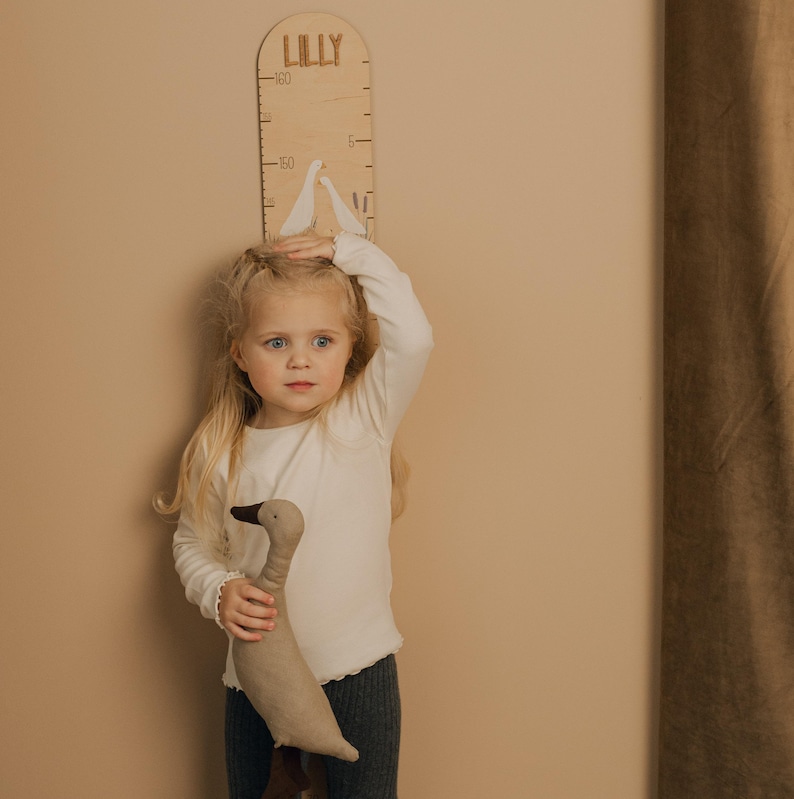 May include: A wooden height chart with the name "LILLY" at the top. The chart has measurement markings and decorative bird cutouts. A young child is standing in front of the chart, holding a stuffed goose toy.