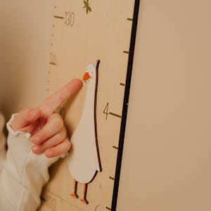 May include: Wooden height chart with a whimsical goose design. The chart features a white goose with orange feet and beak, wearing a party hat. Black measurement markings are visible on the light wood surface. A child's finger points at the goose.