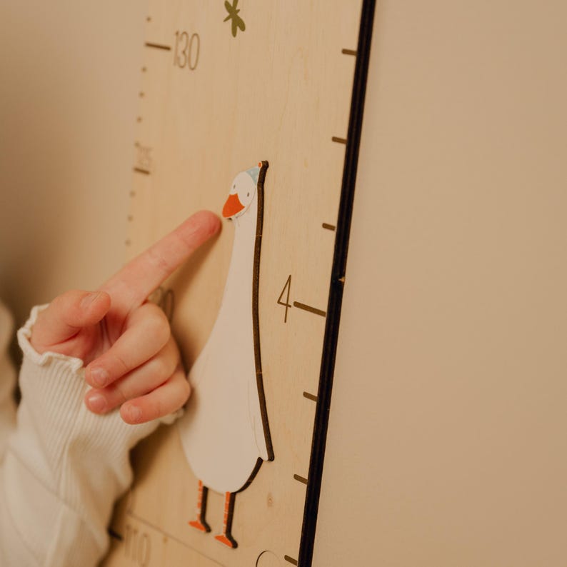 May include: Wooden height chart with a whimsical goose design. The chart features a white goose with orange feet and beak, wearing a party hat. Black measurement markings are visible on the light wood surface. A child's finger points at the goose.