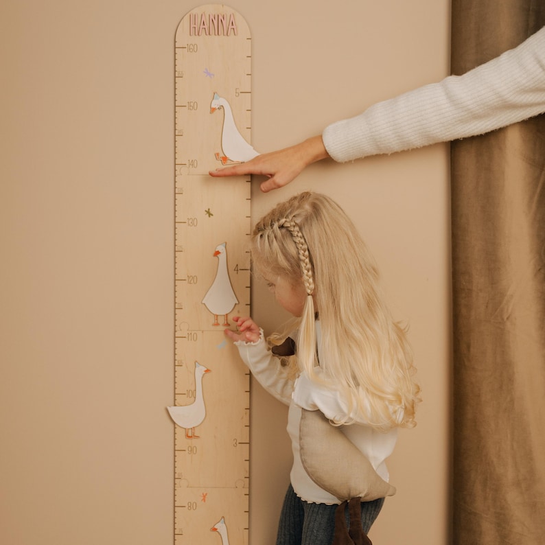 May include: A wooden growth chart with the name "HANNA" at the top. The chart features measurements in inches and decorative white goose cutouts. A child and an adult's hand are interacting with the chart.