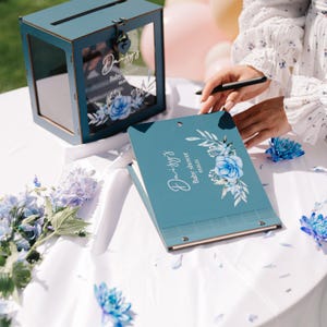 May include: A teal blue guest book with a floral design and the text "Daisy's Baby Shower Wishes" on the cover. The book is open and a person is writing in it. There is a teal blue wooden box with a glass front and a lock on the side. The box has the text "Daisy's Baby Shower Wishes" on the front.