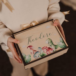 May include: A wooden box with a light brown finish and a dinosaur theme. The box has a hinged lid and is decorated with a watercolor illustration of dinosaurs and greenery. The name "Theodore" is written in cursive on the front of the box.