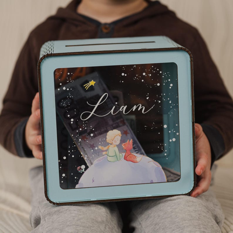 May include: A light blue wooden box with a clear front panel. The box has a slot on the top for inserting money. The front panel features a picture of The Little Prince and the Fox, along with the name "Liam" written in cursive. The box is decorated with a starry night sky.
