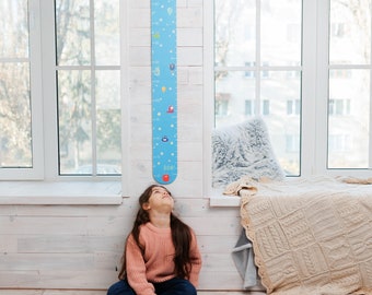 May include: A young girl sits on the floor in front of a blue and white growth chart with colorful illustrations. The chart is mounted on a white wall. The girl is wearing a pink sweater and blue jeans.