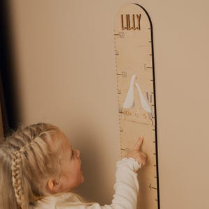 May include: Wooden height chart with the name "LILLY" at the top. The chart features a measurement scale and illustrations of geese. The child is pointing at the chart.