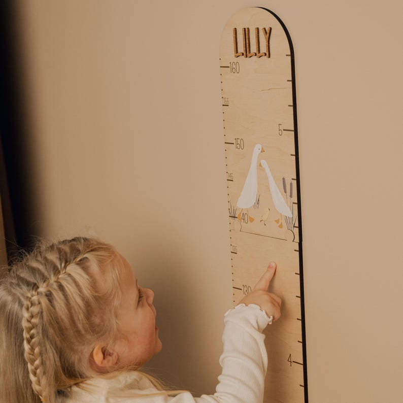 May include: Wooden height chart with the name "LILLY" at the top. The chart features a measurement scale and illustrations of geese. The child is pointing at the chart.