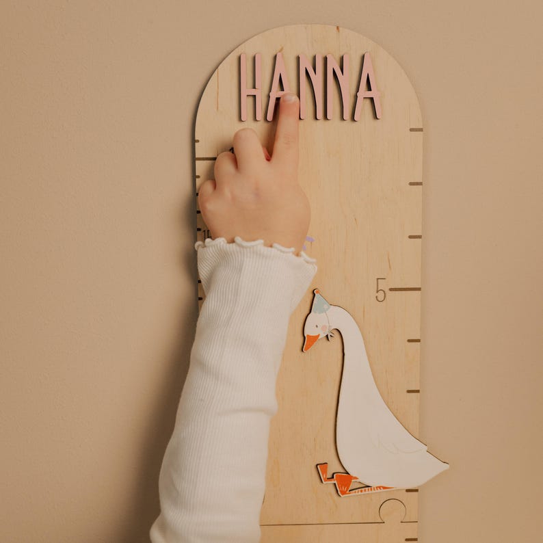 May include: Wooden growth chart with the name "HANNA" in pink letters. A child's hand points to the name. The chart features a decorative goose and measurement markings. The background is a light beige.