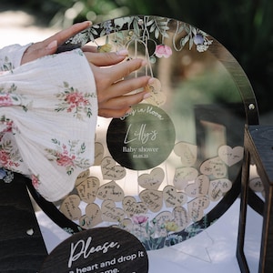 May include: A wooden heart-shaped guest book with a glass front. The guest book is decorated with floral designs and has a sign that says "Baby in Bloom Lilly's Baby Shower 05.05.25". Guests can write messages on small wooden hearts and drop them into the book.