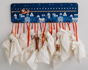 May include: A blue and white wooden advent calendar with 24 small white fabric bags hanging from it. The calendar features a winter village scene with a Santa Claus figure in a sleigh.