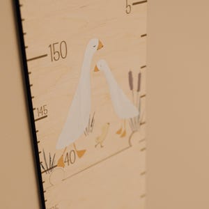 May include: Wooden height chart with a painted illustration of two white geese with orange beaks and feet. The chart has black measurement markings and numbers, with the highest measurement at 150 inches. The background is a light beige color.