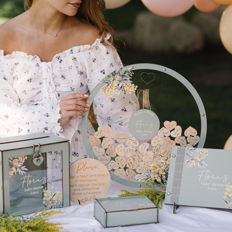 May include: A baby shower guest book with a floral design and the name "Flora's" on the cover. The guest book is open to reveal a blank page. There is also a wooden heart shaped box with a lock and key, a clear round box with wooden hearts inside, and a sign that says "Please sign a heart and drop it in the frame. We'll cherish the love and remember those who came."