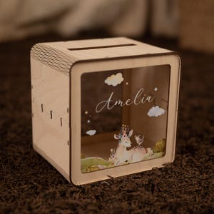 May include: Wooden money box with a clear front panel. The front panel features a whimsical illustration of a deer with a floral crown, a bird, and clouds. The name "Amelia" is written in cursive on the front panel.