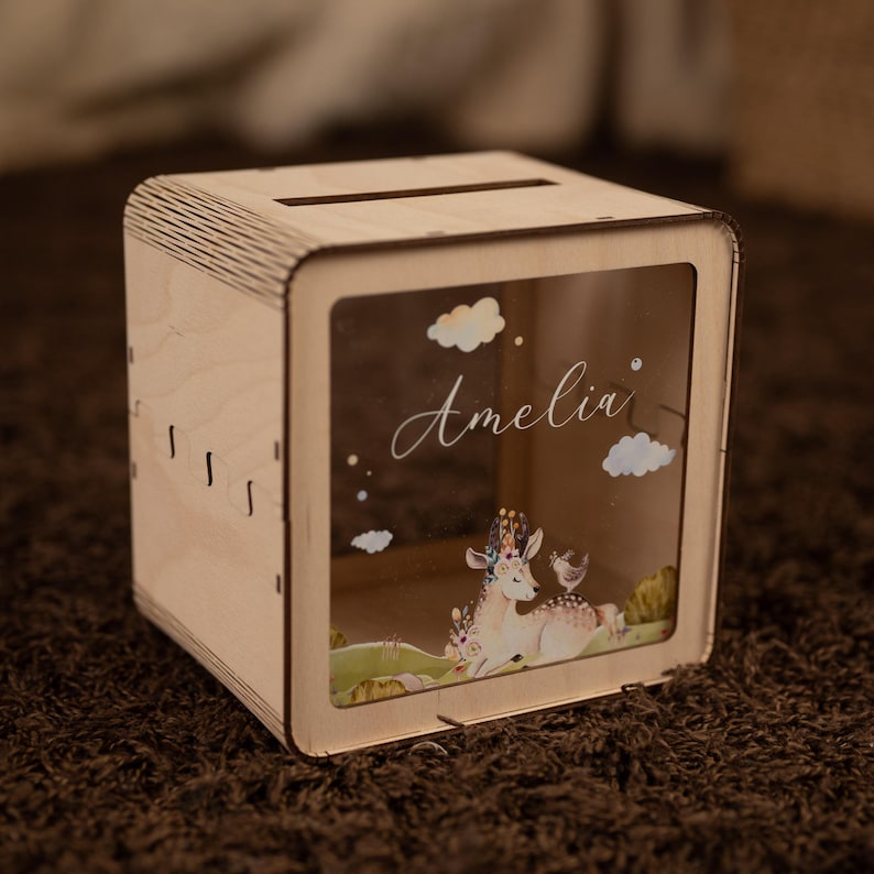 May include: Wooden money box with a clear front panel. The front panel features a whimsical illustration of a deer with a floral crown, a bird, and clouds. The name "Amelia" is written in cursive on the front panel.