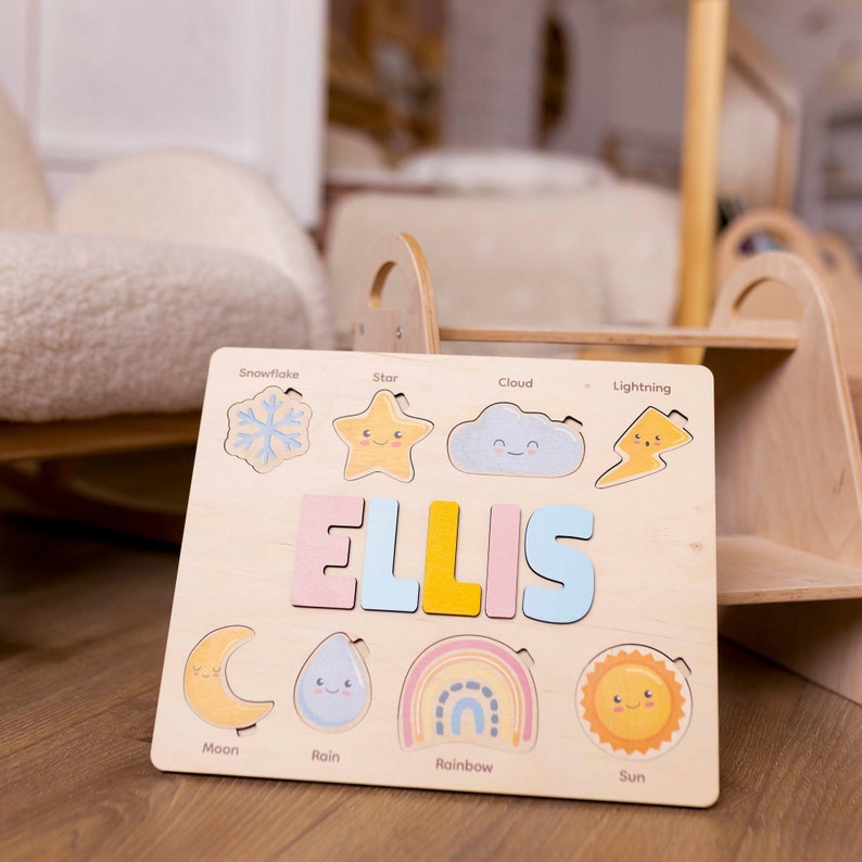 May include: A wooden personalized puzzle with the name "ELLIS" in colorful letters. The puzzle features various weather-themed shapes, including a snowflake, star, cloud, lightning, moon, rain, rainbow, and sun.