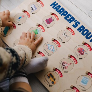 May include: A wooden activity board with a red and white "TO DO" sign and a child's hand pointing to a green recycling bin. The board has a variety of activities for children to complete, including packing a backpack, walking a dog, playing games, painting, going to church, going to school, and drinking water. The board is labeled "HARPER'S ROUTINE".