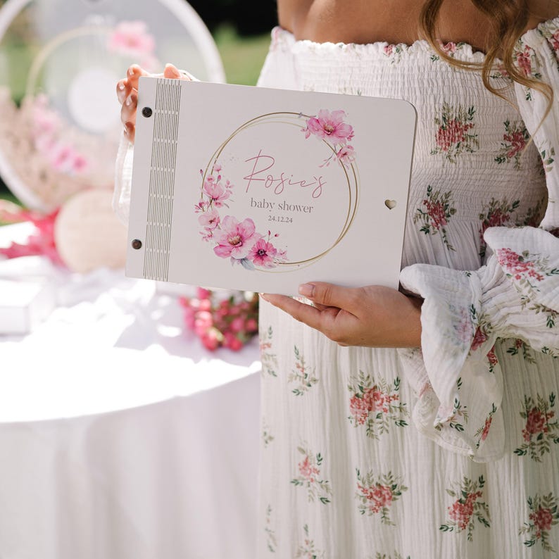May include: A white guest book with a gold circle and pink floral design. The text "Rosie's baby shower 24.2.24" is printed on the front.