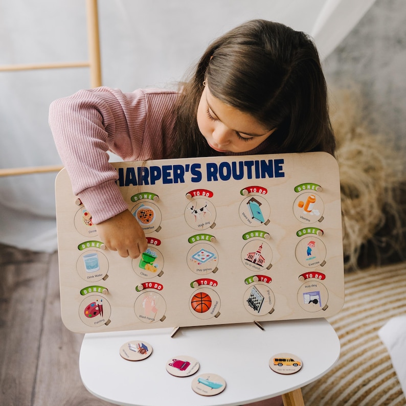 May include: A wooden activity board with the text "Harper's Routine" printed on it. The board has a series of circles with different images and the words "Done" and "To Do" printed on them. A young girl is interacting with the board, moving a small wooden token with an image of a green trash can.