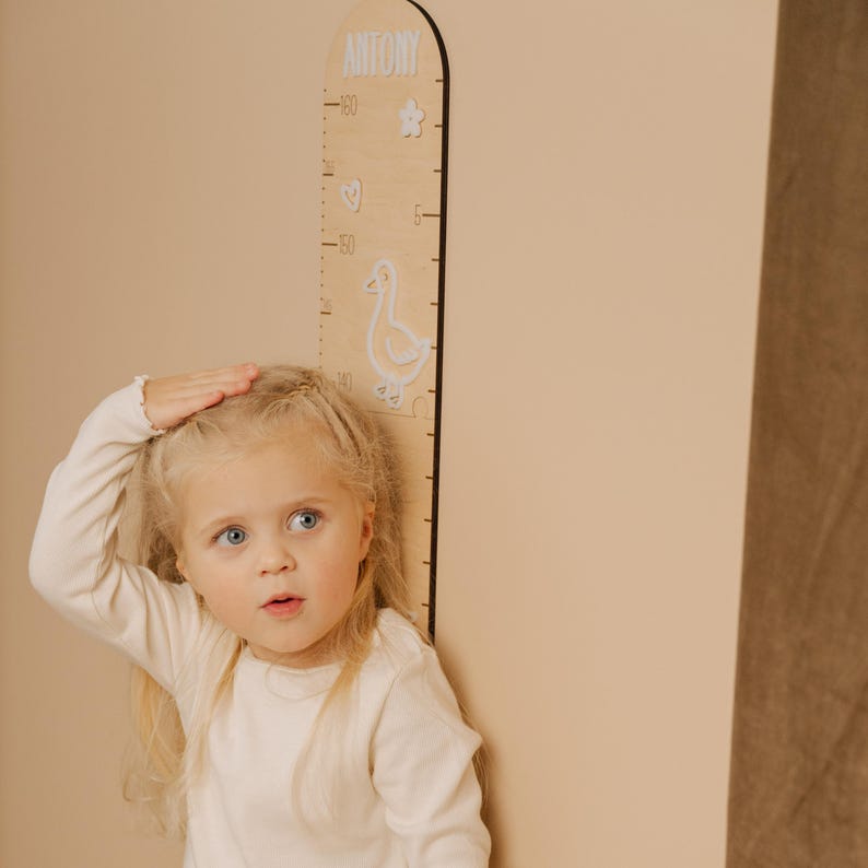 May include: A wooden growth chart with the name "ANTONY" at the top. The chart features a measurement scale, decorative cutouts of a flower, a heart, and a duck. The chart is designed to measure height in inches and centimeters.