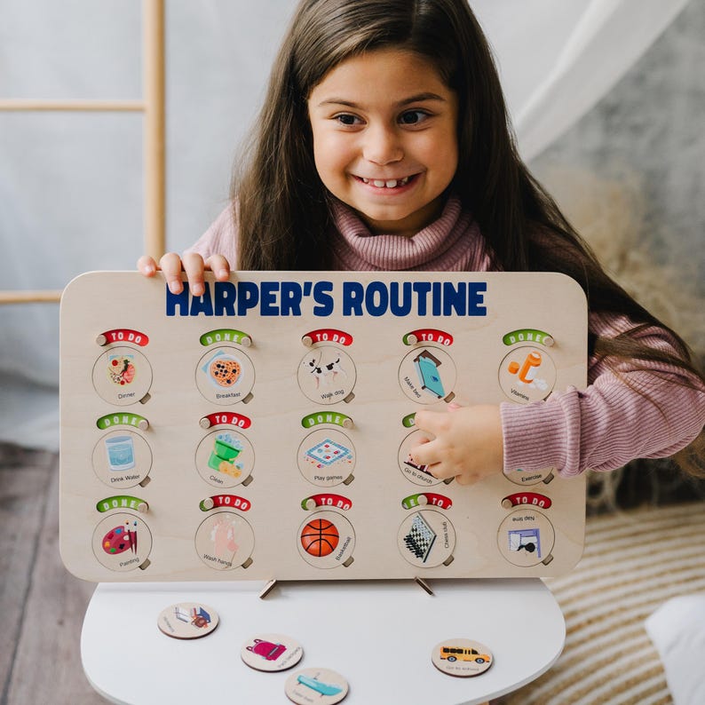 May include: A wooden activity board with the text "HARPER'S ROUTINE" in blue letters. The board has 18 circles with different images representing daily activities, such as eating breakfast, brushing teeth, and playing games. A young girl is interacting with the board, moving a circular token with an image of a school bus.