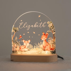 以下が含まれることがあります： 木製のナイトライトで、透明なアクリル製のインサートには、キツネと鹿の森林のシーンが描かれています。アクリル製のインサートには、「エリザベス」という名前が筆記体で書かれています。ナイトライトは下から照らされています。