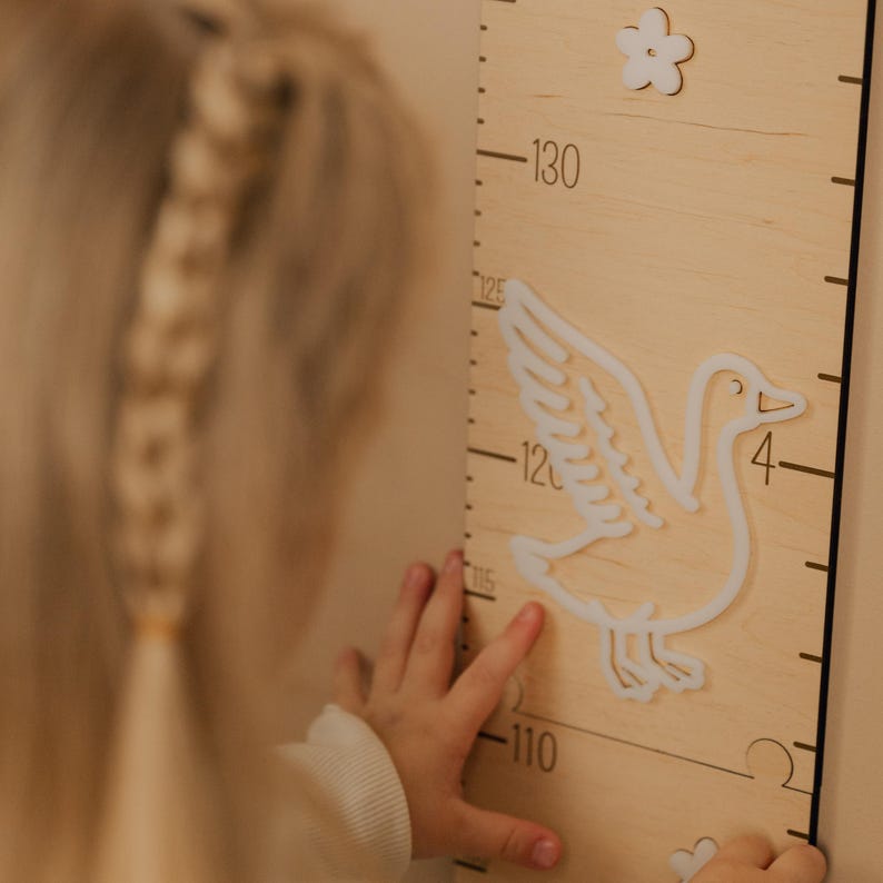 May include: Wooden height chart with a white goose and flower design. The chart has black measurement markings in centimeters, ranging from 110 to 130. A child's hand is reaching towards the chart.