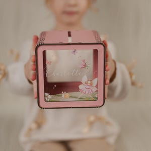 May include: Pink wooden fairy themed money box with a clear front panel. The box has a slot on the top for inserting money. The front panel features a fairy illustration and the name "Charlotte".