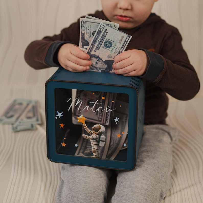 May include: A blue wooden piggy bank with a clear front panel. The front panel features a cartoon astronaut holding a star and a background of stars and dollar bills. The piggy bank is personalized with the name "Mateo".