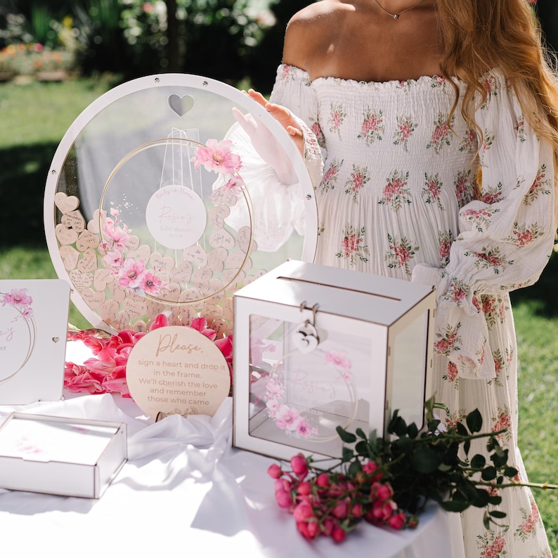 May include: A white wooden box with a glass front and a lock. The box is filled with pink flowers and hearts. There is a sign that says "Please sign a heart and drop it in the frame. We'll cherish the love and remember those who came."