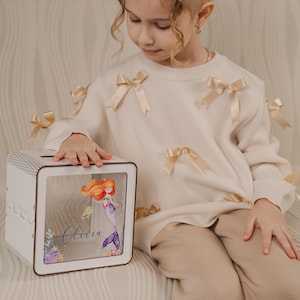May include: White wooden piggy bank with a clear front panel featuring a mermaid illustration and the name Olivia. The piggy bank is sitting on a light brown surface.