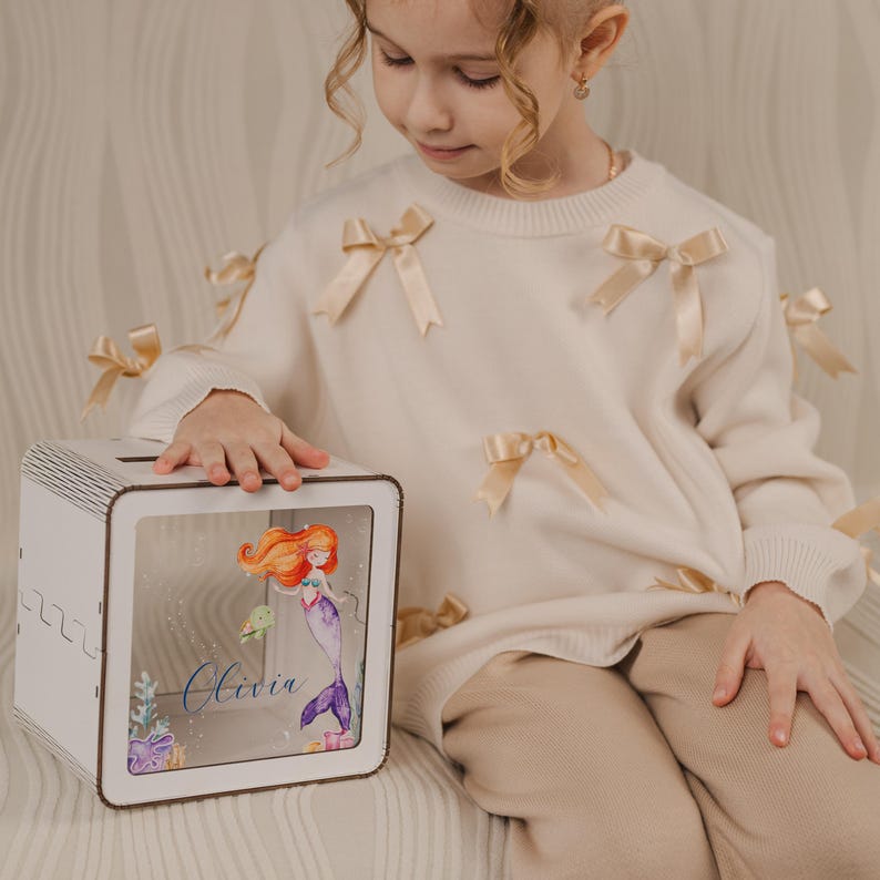 May include: White wooden piggy bank with a clear front panel featuring a mermaid illustration and the name Olivia. The piggy bank is sitting on a light brown surface.