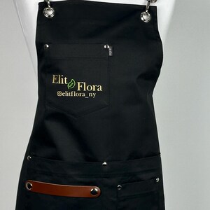 May include: Black apron with brown leather straps and accents. The apron has a pocket with a zipper and two lower pockets. The text "Elit Flora" and "@elitflora_ny" is printed on the apron.