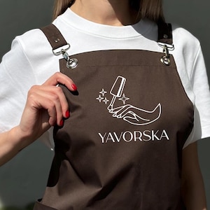 Pode incluir: Avental marrom com um logotipo branco com um design de frasco de esmalte e mão, com o texto "YAVORSKA". O avental tem dois bolsos frontais e alças ajustáveis. A modelo está vestindo uma camiseta branca.