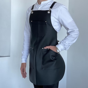 Peut inclure: Tablier en cuir noir avec deux grandes poches avant et des bretelles réglables. Le tablier a une quincaillerie argentée et est porté sur une chemise blanche boutonnée. Conçu pour une utilisation en cuisine ou en atelier.