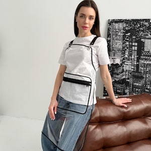 Puede incluir: Delantal transparente con ribetes y correas negras, con un bolsillo en el pecho con cremallera negra. El delantal se lleva sobre una camiseta blanca y unos vaqueros azules. El delantal es rectangular y protege la ropa.