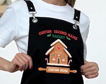 Op de afbeelding: Zwarte schort met verstelbare bandjes en een peperkoekhuisontwerp. De schort heeft de tekst "CUSTOM SECOND NAME BAKERY" en "CUSTOM NAME". Het peperkoekhuis heeft hart- en sterdetails.