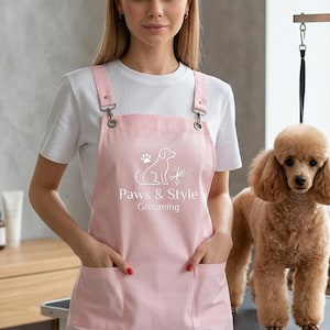 Puede incluir: Un delantal rosa claro con un gráfico blanco de la silueta de un perro, una huella y unas tijeras, con el texto Paws & Style Grooming impreso en blanco. El delantal tiene dos bolsillos delanteros y herrajes metálicos para los tirantes.