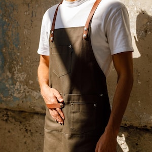 Può includere: Un grembiule marrone scuro con cinghie e tasche in pelle marrone. Il grembiule è indossato sopra una maglietta bianca. Il grembiule ha due tasche ed è fissato con rivetti metallici. Lo sfondo è un muro strutturato di colore neutro.