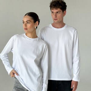 May include: Two people wearing white long-sleeve shirts. The shirts have a crew neck and a simple, clean design. One person is wearing gray jeans, and the other is wearing black jeans. The background is a plain, light gray.