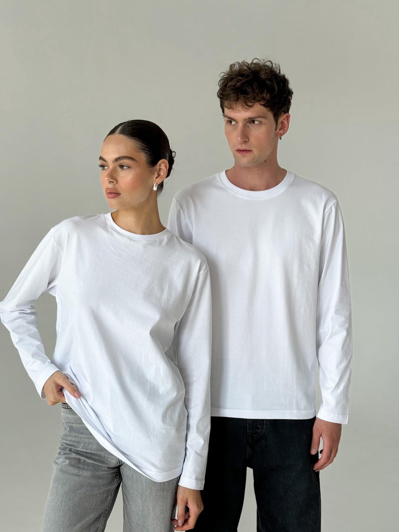 May include: Two people wearing white long-sleeve shirts. The shirts have a crew neck and a simple, clean design. One person is wearing gray jeans, and the other is wearing black jeans. The background is a plain, light gray.